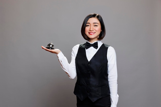 Kostenloses Foto restaurant-rezeptionistin in uniform hält eine glocke in der hand, lächelt und blickt in die kamera. fröhliche junge asiatische kellnerin des catering-service serviert und erregt aufmerksamkeitsporträt