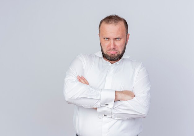 Ärgerlicher bärtiger Mann, der weißes Hemd mit ernstem Gesicht mit verschränkten Armen trägt, die über weißer Wand stehen