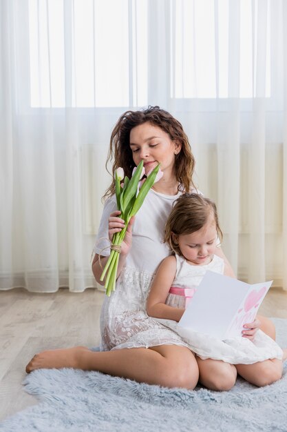 Riechende Blumen der jungen Mutter während kleine Tochterlesegrußkarte zu Hause