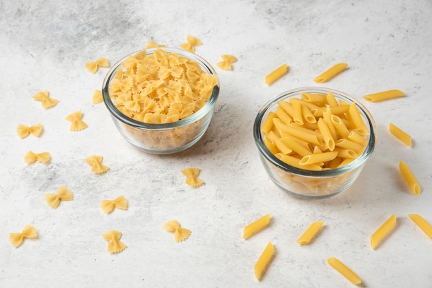 Rohe Farfalle- und Penne-Nudeln in Keramikschalen auf weißer Oberfläche.