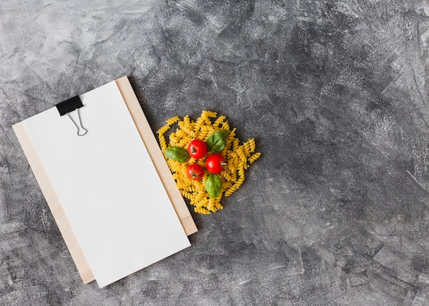 Rohe Fusilli mit Tomaten und Basilikum verlässt mit leerem Papier auf Klemmbrett gegen strukturierten Hintergrund