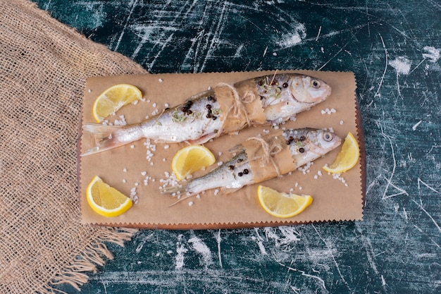 Rohe ganze Fische mit Zitronenscheiben, Pfefferkörnern und Salz auf Holzbrett.