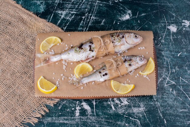 Rohe ganze Fische mit Zitronenscheiben, Pfefferkörnern und Salz auf Holzbrett.