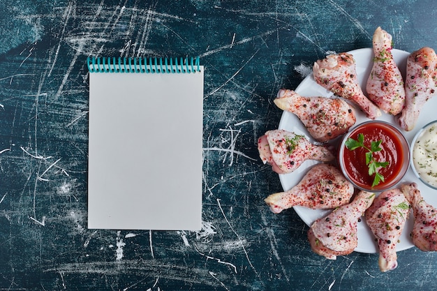 Rohe Hähnchenschenkel auf einer weißen Platte mit einem Rezeptbuch beiseite.