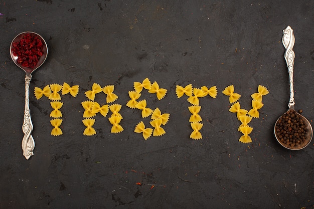 Kostenloses Foto rohe pasta leckeres wort geformt gelb zusammen mit zwei silbernen löffeln mit verschiedenen gewürzen auf einem dunklen schreibtisch