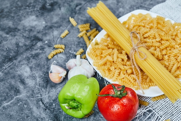 Rohe Spaghetti und Fusilli auf einem weißen Teller mit Tomaten, Pfeffer und Knoblauch.