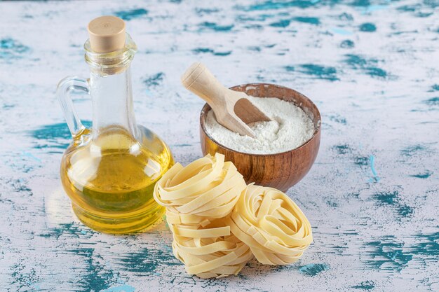 Rohe Tagliatelle-Nester, Olivenöl und Mehlschale auf bunter Oberfläche.