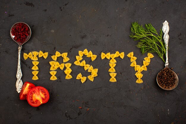 Rohes leckeres Wort der gelben Nudeln zusammen mit geschnittenen Tomaten und Grüns auf einem grauen Boden geformt