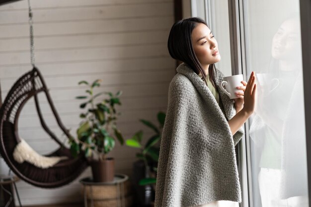 Romantische asiatische Frau, die das Fenster berührt und nach draußen schaut, mit nachdenklichem Gesicht, das Tee trinkt und keine...