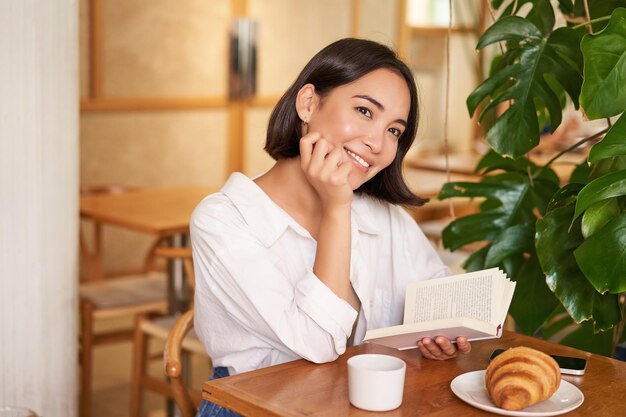 Romantische asiatische Frau, die mit Buch im Café sitzt und Croissants isst und Kaffee trinkt, liest und lächelt