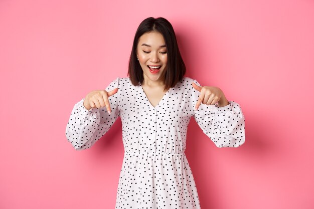 Romantische asiatische Frau, die nach unten schaut, mit den Fingern nach unten zeigt, neugierig auf den Produktrabatt mit glücklichem Lächeln schaut und über rosafarbenem Hintergrund steht