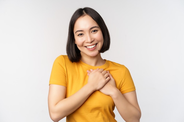 Romantische asiatische Freundin, die Hände am Herzen hält und mit Sorgfalt und Zärtlichkeit lächelt und in gelbem T-Shirt auf weißem Hintergrund steht