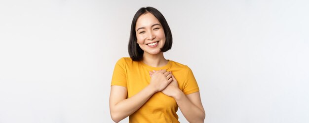 Romantische asiatische Freundin, die Hände am Herzen hält und mit Sorgfalt und Zärtlichkeit lächelt und in gelbem T-Shirt auf weißem Hintergrund steht