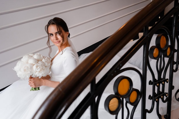 Romantische Braut in klassischem Satinkleid mit Blumenstrauß aus Pionies, die sich auf der Treppe im Innenbereich rühren Frau hat eine schöne Hochzeitsfrisur mit Schleier-Accessoires und nacktem natürlichem Make-up