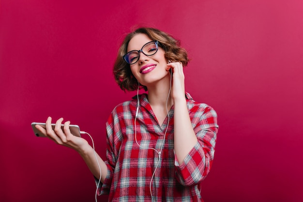 Romantische Frau mit erfreutem Lächeln, das Musik in den Kopfhörern hört. Innenporträt des fröhlichen weißen weiblichen Modells in den Gläsern mit der kurzen Frisur, die auf Rotweinwand steht.