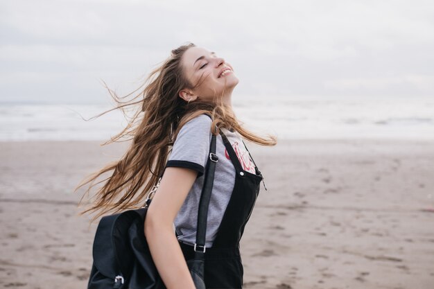 Romantische langhaarige Frau mit schwarzem Rucksack, die guten Tag am Sandstrand genießt. Außenaufnahme des kaukasischen reizenden Mädchens springend.