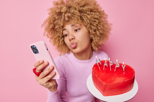 Romantische, lockige junge Frau hält die Lippen gefaltet, macht Selfie per Smartphone und hält köstlichen herzförmigen Kuchen, der sich auf den Valentinstag vorbereitet, gekleidet in einem Pullover, der isoliert über rosa Hintergrund gekleidet ist