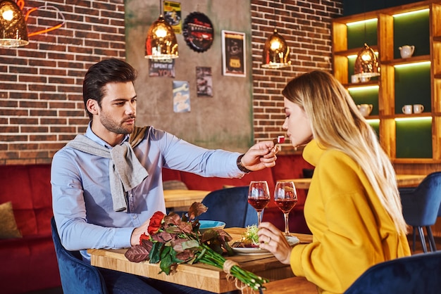 Romantischer dinner-mann im blauen hemd füttert seine hübsche blonde freundin