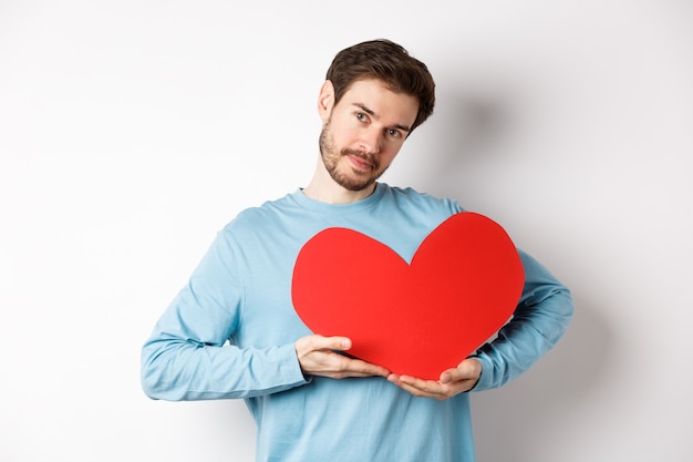 Romantischer Freund, der die Valentinstagsüberraschung macht, einen großen roten Herzausschnitt auf der Brust hält und mit Liebe lächelt, zärtlich in die Kamera schaut und auf weißem Hintergrund steht