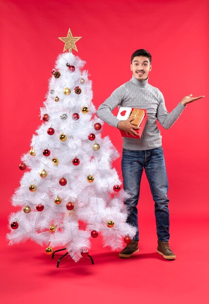 Romantischer hübscher Erwachsener in einer grauen Bluse, die nahe dem geschmückten weißen Weihnachtsbaum steht