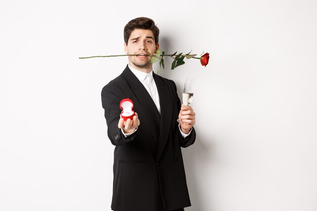 Romantischer junger Mann im Anzug, der einen Vorschlag macht, eine Rose in den Zähnen und ein Glas Champagner hält, einen Verlobungsring zeigt, ihn um eine Heirat bittet und vor weißem Hintergrund steht