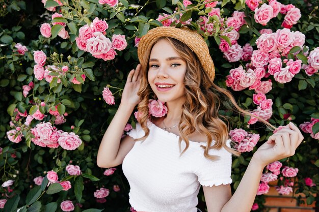 Romantisches blondes Mädchen, das mit Lächeln vor schönen Blumen aufwirft. Außenporträt der fröhlichen lockigen Frau, die mit ihren Haaren im Garten spielt.