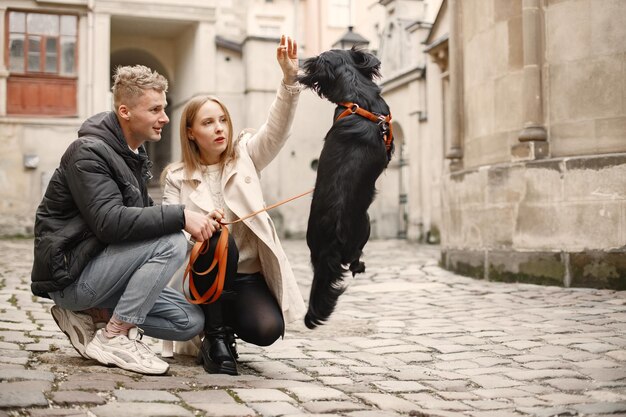 Romantisches Paar steht auf der Straße im Herbst Mann und Frau streicheln einen schwarzen Hund auf einer Straße in der Altstadt Mädchen mit beigem Mantel und schwarzer Jacke des Mannes