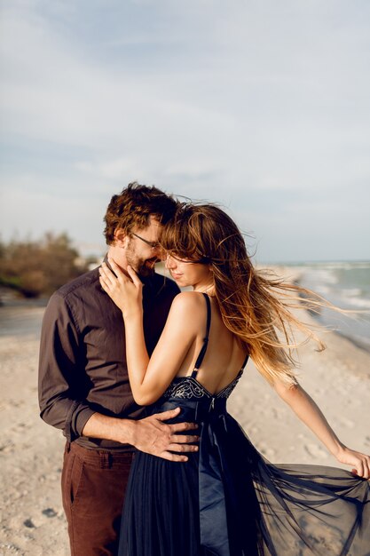 Romantisches Paar umarmen am Abendstrand nahe Meer. Elegante Frau im blauen Kleid, die ihren Freund mit Zärtlichkeit umarmt.