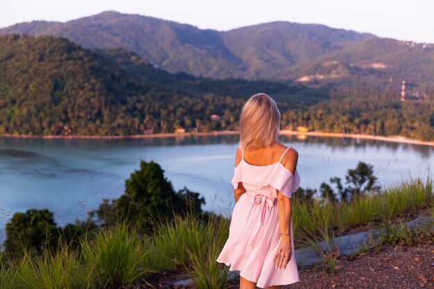 Romantisches Porträt der jungen kaukasischen Frau im Sommerkleid, das das Entspannen im Park auf Berg mit erstaunlichem tropischem Meerblick genießt Frau auf Urlaubsreise durch Thailand Glückliche Frau bei Sonnenuntergang