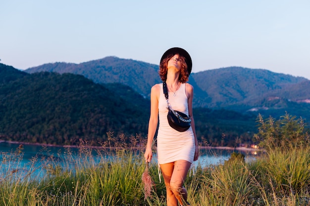 Romantisches Porträt der jungen kaukasischen Frau im Sommerkleid, das das Entspannen im Park auf Berg mit erstaunlichem tropischem Meerblick genießt Frau auf Urlaubsreise durch Thailand Glückliche Frau bei Sonnenuntergang