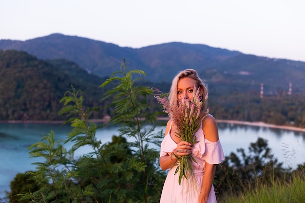 Romantisches Porträt der jungen kaukasischen Frau im Sommerkleid, das das Entspannen im Park auf Berg mit erstaunlichem tropischem Meerblick genießt
