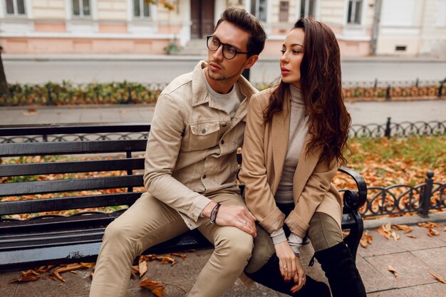 Romantisches Porträt des jungen schönen Paares in der Liebe, die auf Bank im Herbstpark umarmt und küsst. Trage einen stilvollen beigen Mantel.