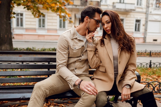 Romantisches Porträt des jungen schönen Paares in der Liebe, die auf Bank im Herbstpark umarmt und küsst. Trage einen stilvollen beigen Mantel.