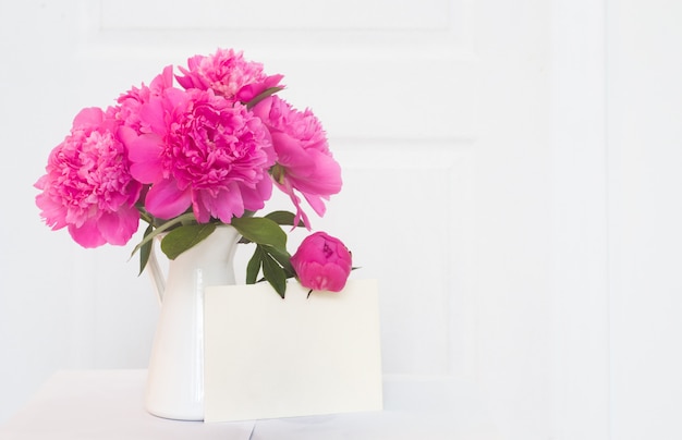 Rosa Pfingstrosen in weißer emaillierter Vase. Schöne Blumen im Innendesign. Weißes Papier für Einladungstext, weiße Pfingstrosen in einer Vase, Innendekoration