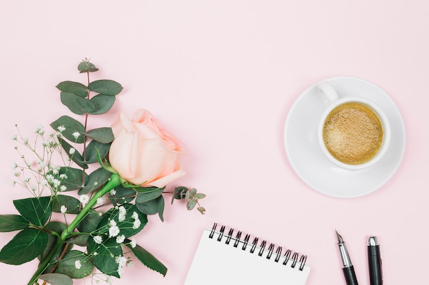 Rosenblume mit Kaffee; Spiralblock und Füllfederhalter auf rosa Hintergrund