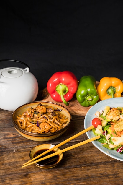 Kostenloses Foto rot; grüne und gelbe paprikaschoten; teekanne; udon-nudeln und salat mit sojasaucenschüssel mit essstäbchen auf dem schreibtisch