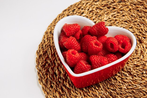 Rote Himbeeren in einer Platte in der Form der Herz-Hochwinkelansicht auf einem Weiden-Tischset und weißem Hintergrund