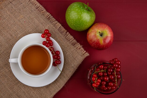 Rote johannisbeere der draufsicht in einem glas mit einer tasse tee und äpfeln auf einem roten hintergrund