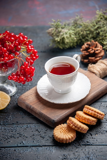 Rote Johannisbeere der unteren Ansicht in einem Glas eine Tasse Tee auf Schneidebrettscheibe Zitronen-Tannenzapfen und Kekse auf dunklem Holzhintergrund