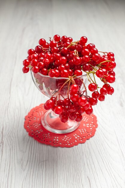 Rote Johannisbeere der Vorderansicht in einem Kristallglas auf dem roten ovalen Spitzendeckchen auf dem weißen Holztisch mit Kopienraum