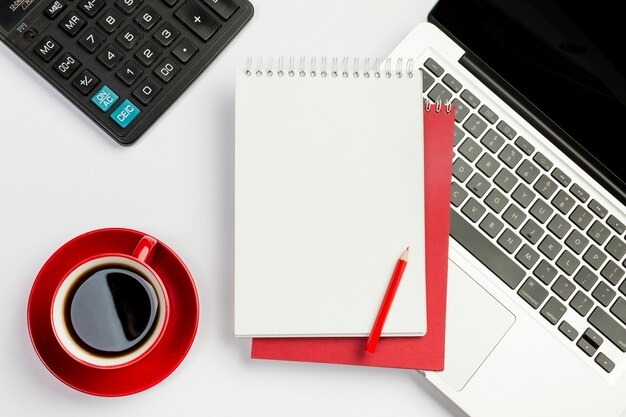 Rote Kaffeetasse, Taschenrechner, gewundener Notizblock, Bleistift auf Laptop über dem weißen Hintergrund