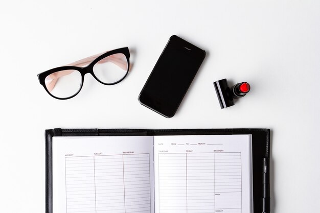 Rote Lippenstift-Telefonbrille und Notizbuch über weißer Oberfläche oben.