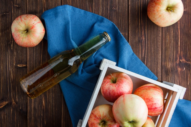 Rote Äpfel in einer Holzkiste und herum mit Apfelsaft-Draufsicht auf einem blauen Tuch und einem hölzernen Hintergrund