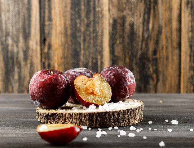 Rote Pflaumen mit Salz auf einem Holzstück auf hölzerner und grauer Oberfläche, Seitenansicht.