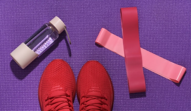 Rote sportschuhe, fitness-gummibänder, eine flasche wasser auf einer lila sportmatte. trainingskonzept.