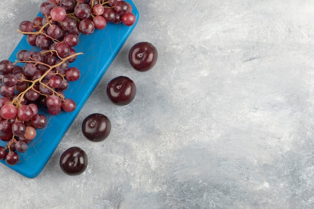 Rote trauben auf blauem schneidebrett mit frischen pflaumen auf marmoroberfläche.