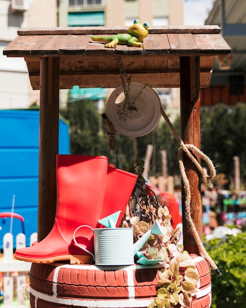 Roter Gummistiefel; Gießkanne und Spaten über den handgefertigten Brunnen im Garten