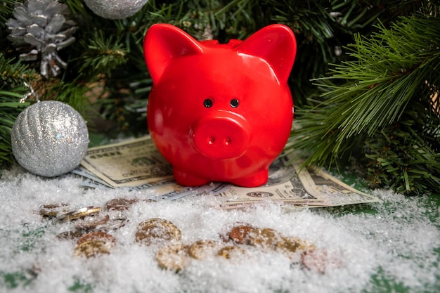 Rotes sparschwein auf goldmünzen und papiergeld bedeckt mit schnee und weihnachtlichen grünen fichtenzweigen, die mit kugeln verziert werden. erfolgskonzept. sparkonzept. vermögenskonzept im neuen jahr