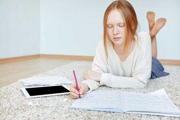 Rothaarige Frau, die auf Boden mit Notizbuch und Tablette liegt