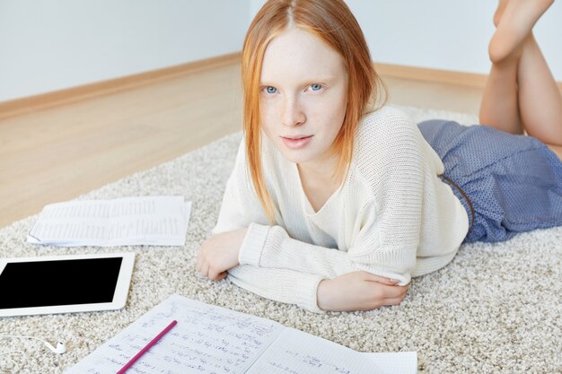 Rothaarige Frau, die auf Boden mit Notizbuch und Tablette liegt